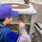 A technician in a blue uniform and cap repairs an outdoor air conditioning unit, working with exposed wires and components—a scene common for Boiler Service NYC professionals handling heating and cooling systems. A technician in a blue uniform and cap repairs an outdoor air conditioning unit, working with exposed wires and components—a scene common for Boiler Service NYC professionals handling heating and cooling systems.