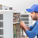 A technician in a blue cap and shirt repairs or inspects the wiring inside an outdoor air conditioning unit, using a screwdriver, with the unit's panel open—just like expert Boiler Service NYC professionals handle heating systems. A technician in a blue cap and shirt repairs or inspects the wiring inside an outdoor air conditioning unit, using a screwdriver, with the unit's panel open—just like expert Boiler Service NYC professionals handle heating systems.