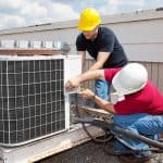 Two workers wearing hard hats inspect and repair a rooftop air conditioning unit on a sunny day. One uses tools while the other observes, showcasing the same expertise they bring to Boiler Service NYC projects. Two workers wearing hard hats inspect and repair a rooftop air conditioning unit on a sunny day. One uses tools while the other observes, showcasing the same expertise they bring to Boiler Service NYC projects.