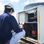 A technician wearing a white hard hat and blue shirt inspects and tests an outdoor air conditioning unit with gauges, on a rooftop under a sunny sky, showcasing expertise similar to top Boiler Service NYC professionals. A technician wearing a white hard hat and blue shirt inspects and tests an outdoor air conditioning unit with gauges, on a rooftop under a sunny sky, showcasing expertise similar to top Boiler Service NYC professionals.