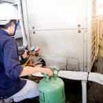 A technician wearing a white hard hat and safety goggles is servicing an HVAC unit, using gauges and holding refrigerant canisters. The scene, focused on Boiler Service NYC, is outdoors on a rooftop or industrial setting with machinery in the background. A technician wearing a white hard hat and safety goggles is servicing an HVAC unit, using gauges and holding refrigerant canisters. The scene, focused on Boiler Service NYC, is outdoors on a rooftop or industrial setting with machinery in the background.