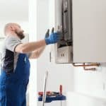 A technician in blue overalls and gloves inspects or repairs a wall-mounted boiler or heating unit in a bright, modern room. Tools are placed on a cart beside him, showcasing professional Boiler Service NYC expertise. A technician in blue overalls and gloves inspects or repairs a wall-mounted boiler or heating unit in a bright, modern room. Tools are placed on a cart beside him, showcasing professional Boiler Service NYC expertise.