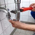 A person uses a red pipe wrench to fix a leaky metal pipe under a sink, with water spraying onto tiled walls and floor—just another busy day for a Boiler Service NYC technician. A person uses a red pipe wrench to fix a leaky metal pipe under a sink, with water spraying onto tiled walls and floor—just another busy day for a Boiler Service NYC technician.