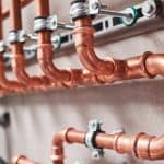 Close-up view of several copper pipes with metal joints and clamps mounted on a wall, forming part of a building’s plumbing or heating system—typical components inspected during Boiler Service NYC. Close-up view of several copper pipes with metal joints and clamps mounted on a wall, forming part of a building’s plumbing or heating system—typical components inspected during Boiler Service NYC.